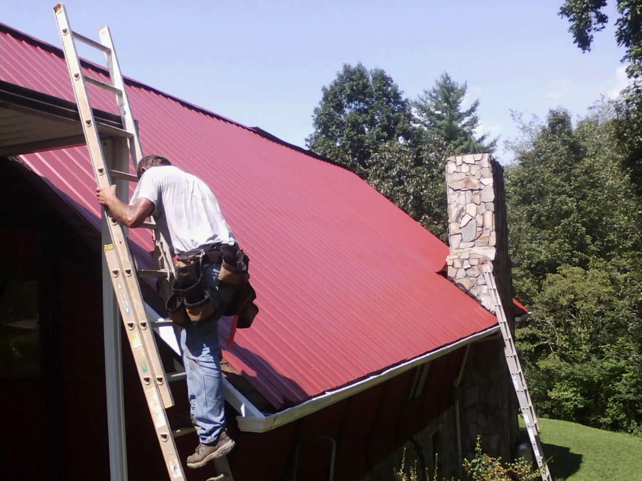 Roofer on a ladder beside a red metal roof and stone chimney — Soffit & Fascia Repair by Roofing Brothers in Ashfield